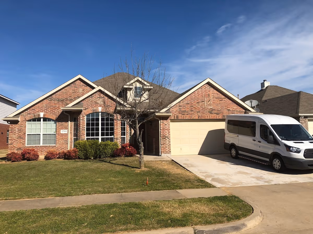A single-story brick house with a two-car garage and a white van parked in the driveway. The house has large windows, a small tree in the front yard, and neatly trimmed bushes. The sky is clear with a few clouds.