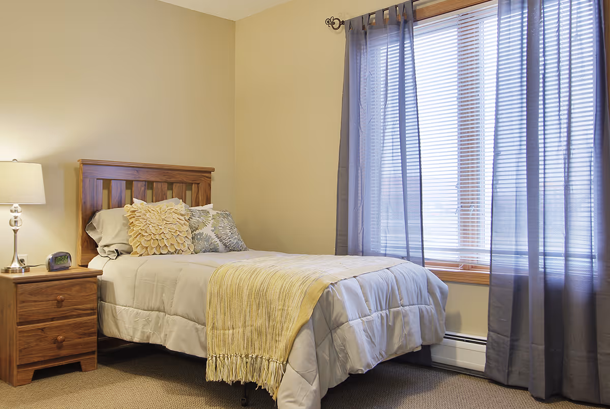 A cozy bedroom with a single wooden bed featuring a beige comforter, decorative pillows, and a yellow throw blanket. Next to the bed is a wooden nightstand with a lamp and a digital alarm clock. A large window with blinds and sheer gray curtains allows natural light into the room.