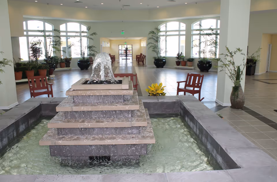 Sunlit indoor lobby with tiered fountain, benches, and numerous potted plants.
