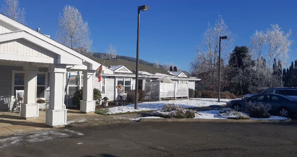 Front exterior of a single-story senior living building with a covered porch, small snow-covered lawn, lamp posts and parked cars.