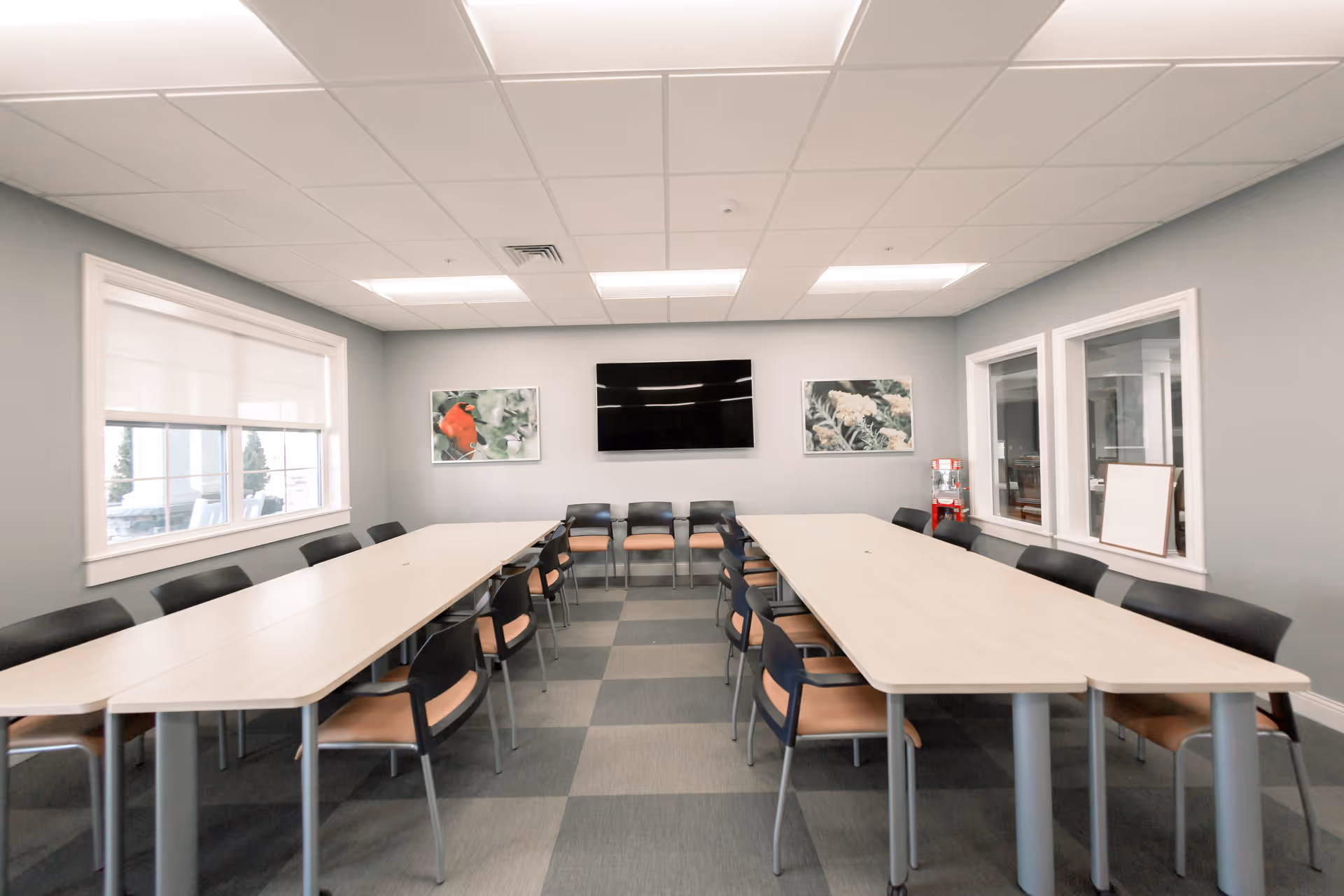 A conference or meeting room with two long tables arranged in a U-shape, surrounded by chairs. The room has a large flat-screen TV mounted on the wall, two framed pictures of flowers, a window with blinds, and a water dispenser in the corner. The floor has a checkered pattern carpet, and the walls are painted light gray.