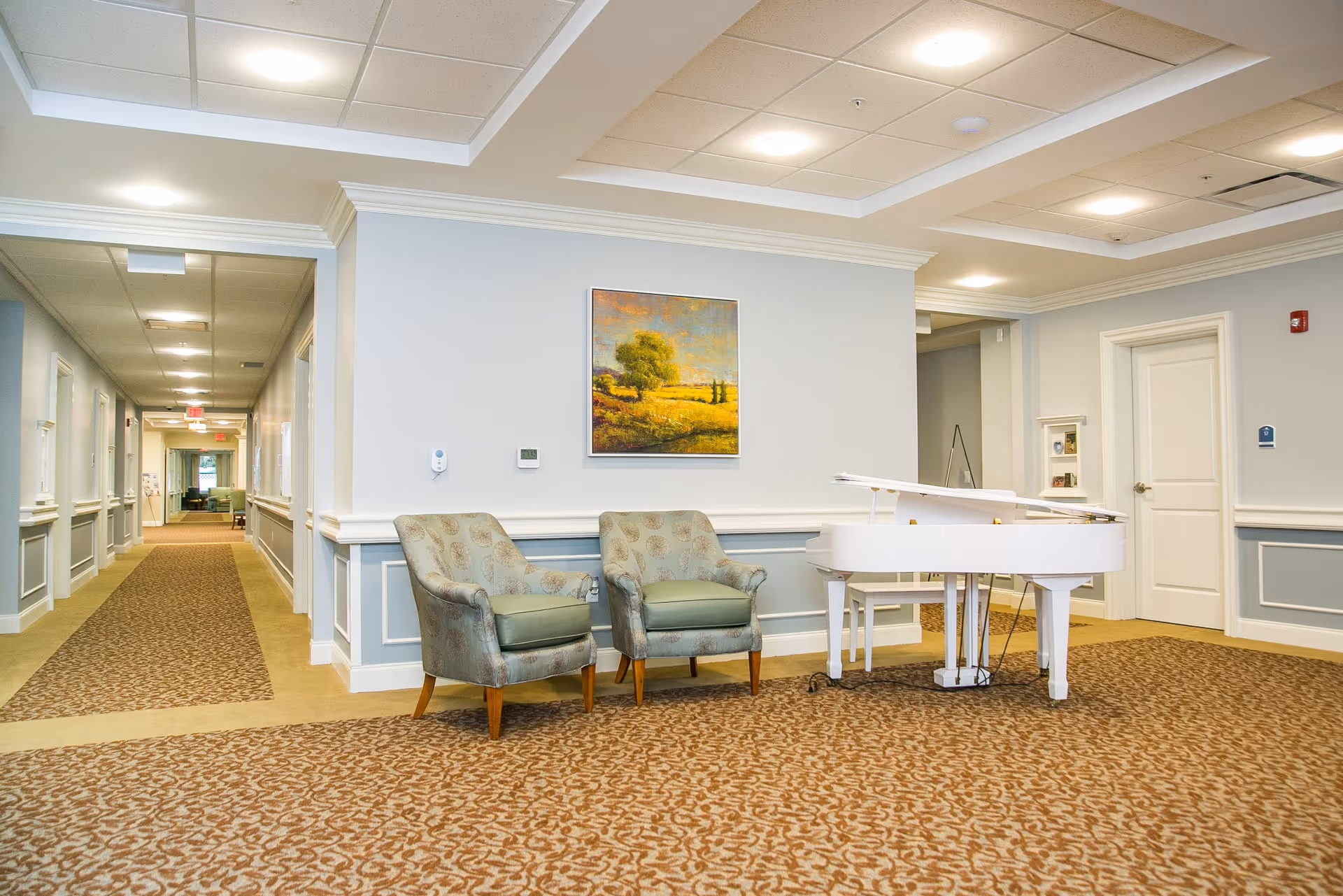 Interior hallway of a senior living facility with patterned carpet, two upholstered armchairs, a white grand piano, and a landscape painting on the wall. The hallway extends into the distance with doors and lighting fixtures along the walls.