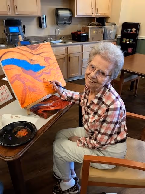 An elderly woman wearing glasses and a plaid shirt is sitting at a table in a room with wooden cabinets and a sink. She is painting on a canvas with bright orange, blue, and purple colors and smiling at the camera. The room appears to be a communal or activity area with art supplies on the table.