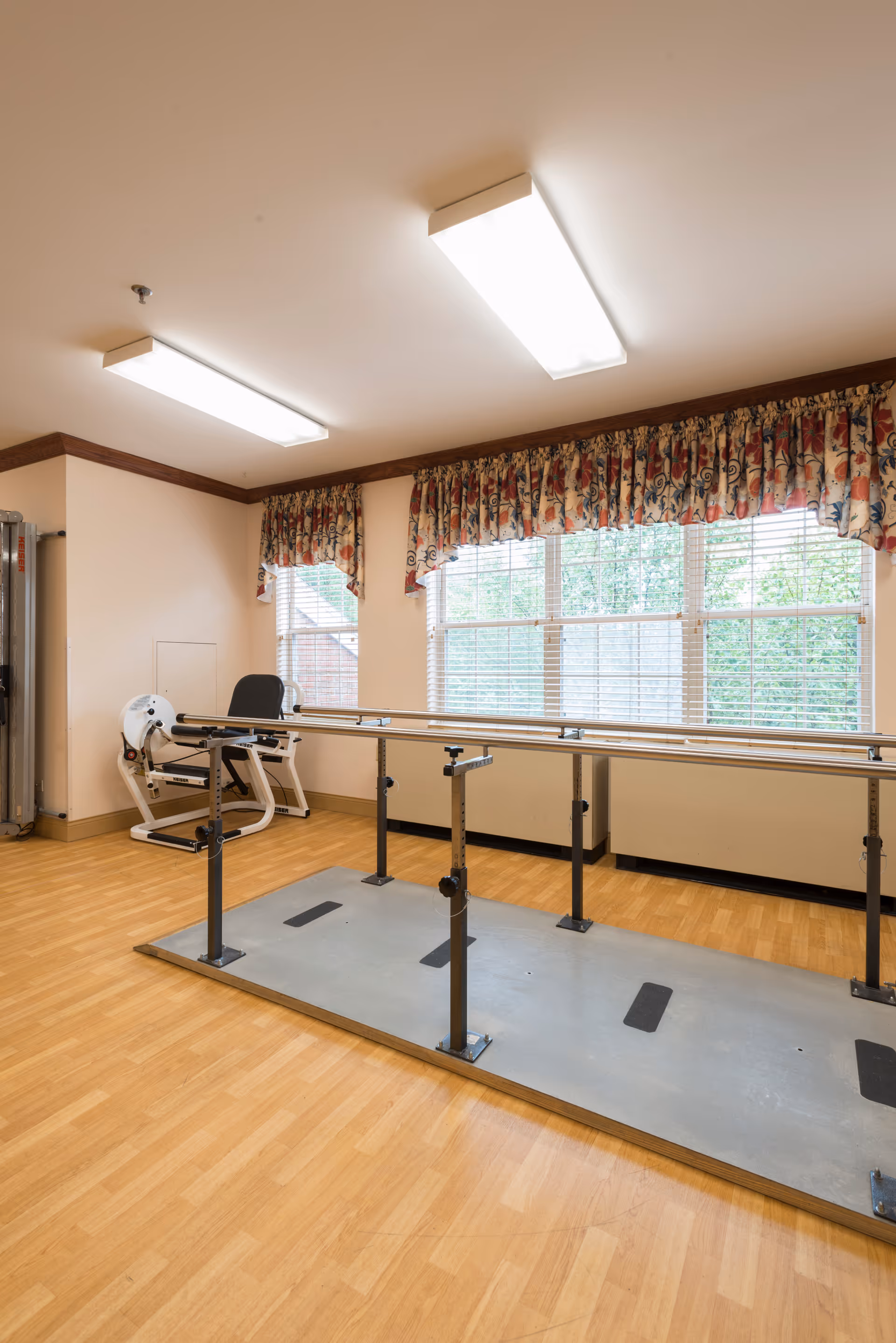A rehabilitation or physical therapy room with parallel bars mounted on a platform for walking exercises, a leg exercise machine, large windows with floral valance curtains, and wood-patterned flooring.
