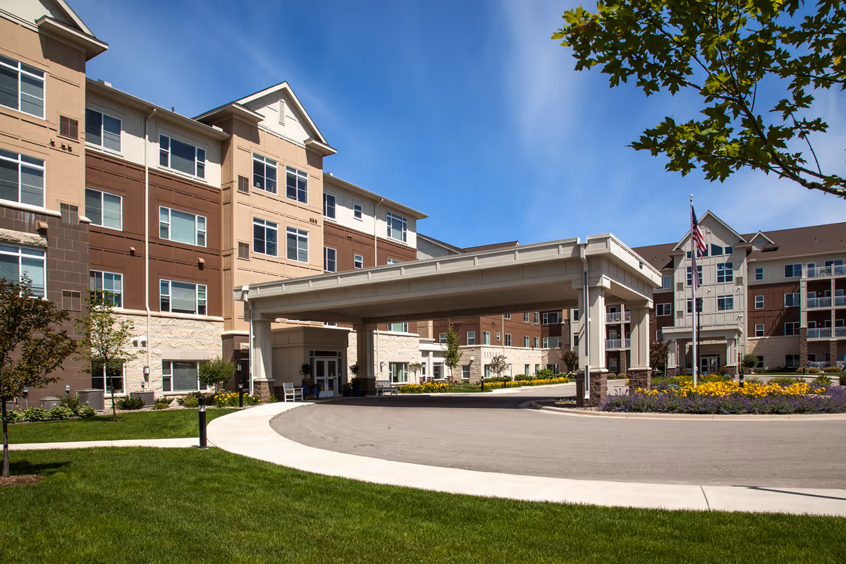 Exterior view of a multi-story senior living facility with a covered entrance driveway, well-maintained landscaping including green grass, trees, and flower beds, and an American flag on a flagpole under a clear blue sky.