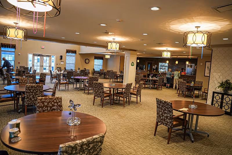 Spacious senior living dining area with round tables, patterned chairs, hanging light fixtures and a small serving bar in the background.