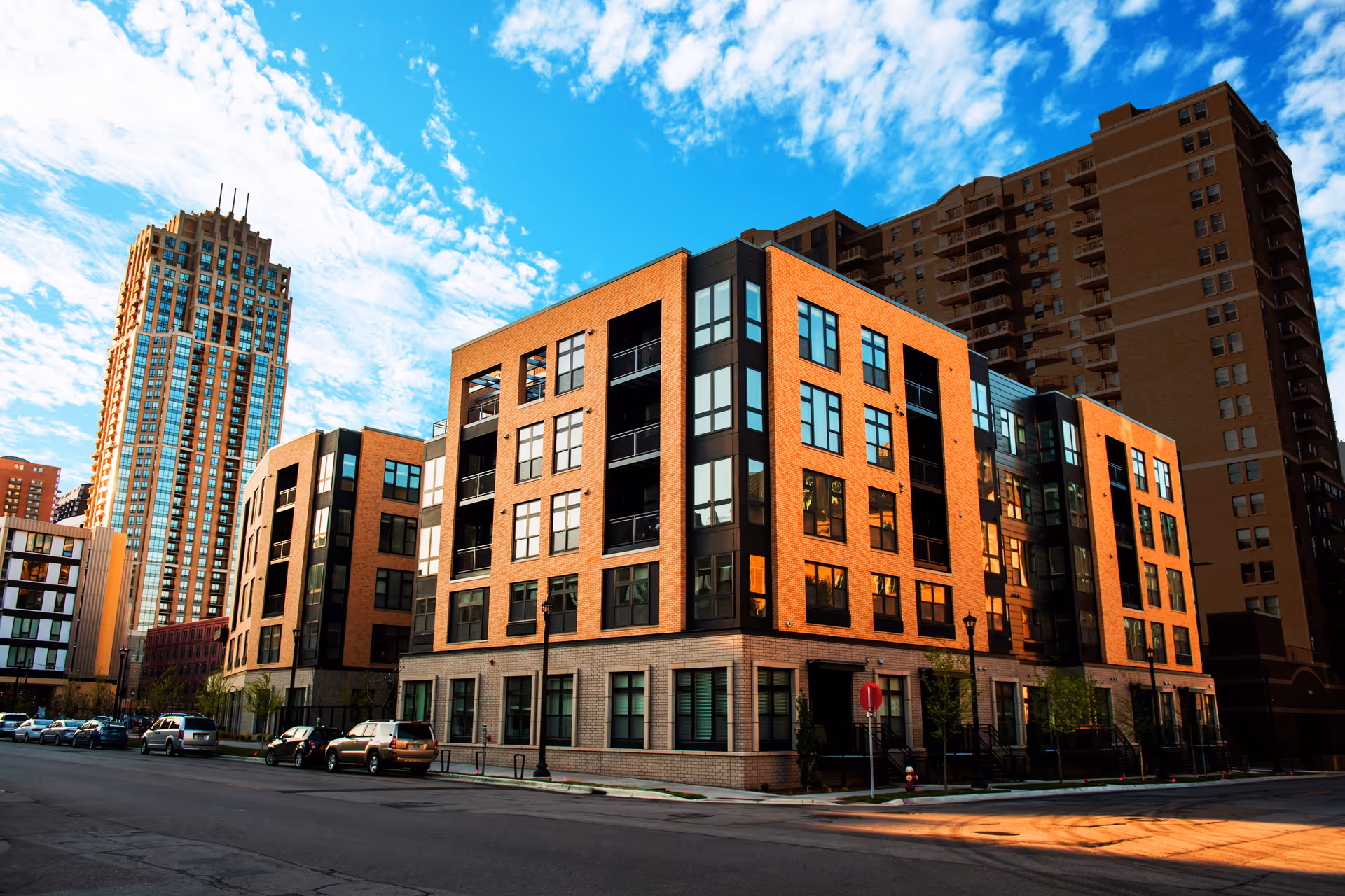 Modern multi-story residential building with large windows and balconies, situated on a city street with parked cars and other tall buildings in the background under a partly cloudy blue sky.