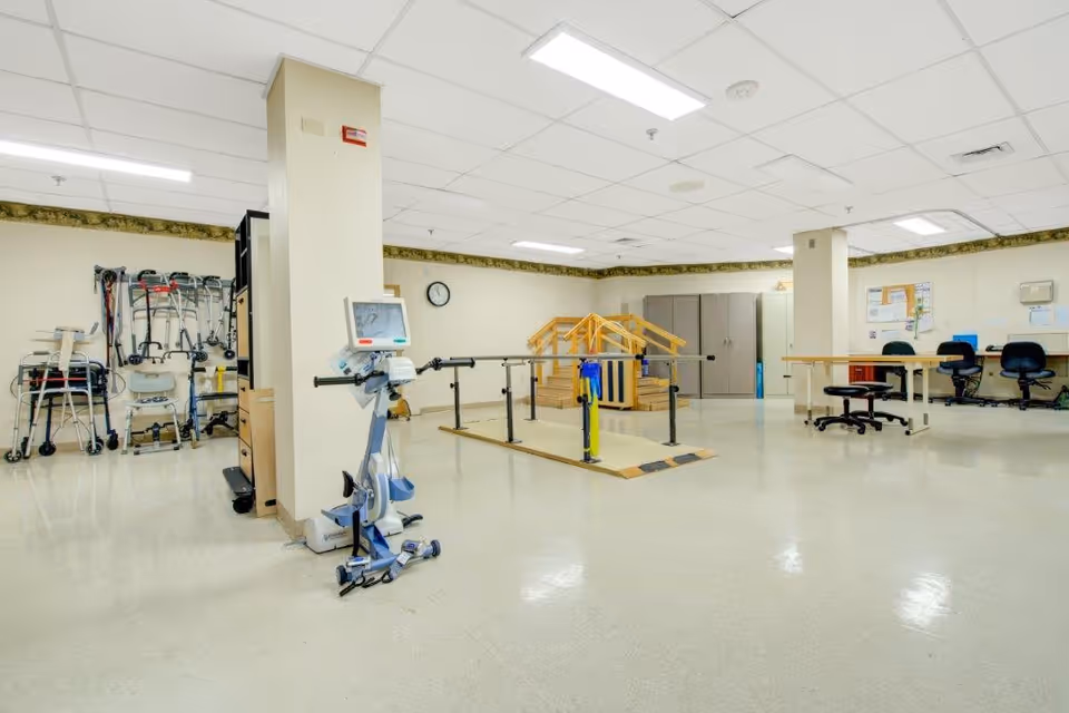 A spacious rehabilitation room with parallel bars and a small set of stairs for physical therapy exercises. The room also contains various walkers and mobility aids lined up against the wall, a medical device on wheels, desks with chairs, and cabinets. The ceiling has fluorescent lighting and the floor is a polished light-colored surface.