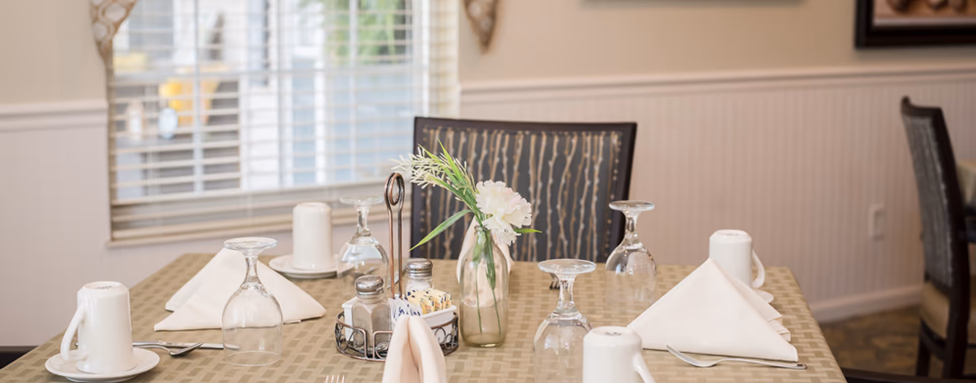 A dining table set for four with upside-down wine glasses, white folded napkins, white coffee cups on saucers, silverware, a small vase with white flowers and greenery, and a condiment holder with salt, pepper, and sugar packets. The background shows a window with blinds and part of the dining room with chairs.