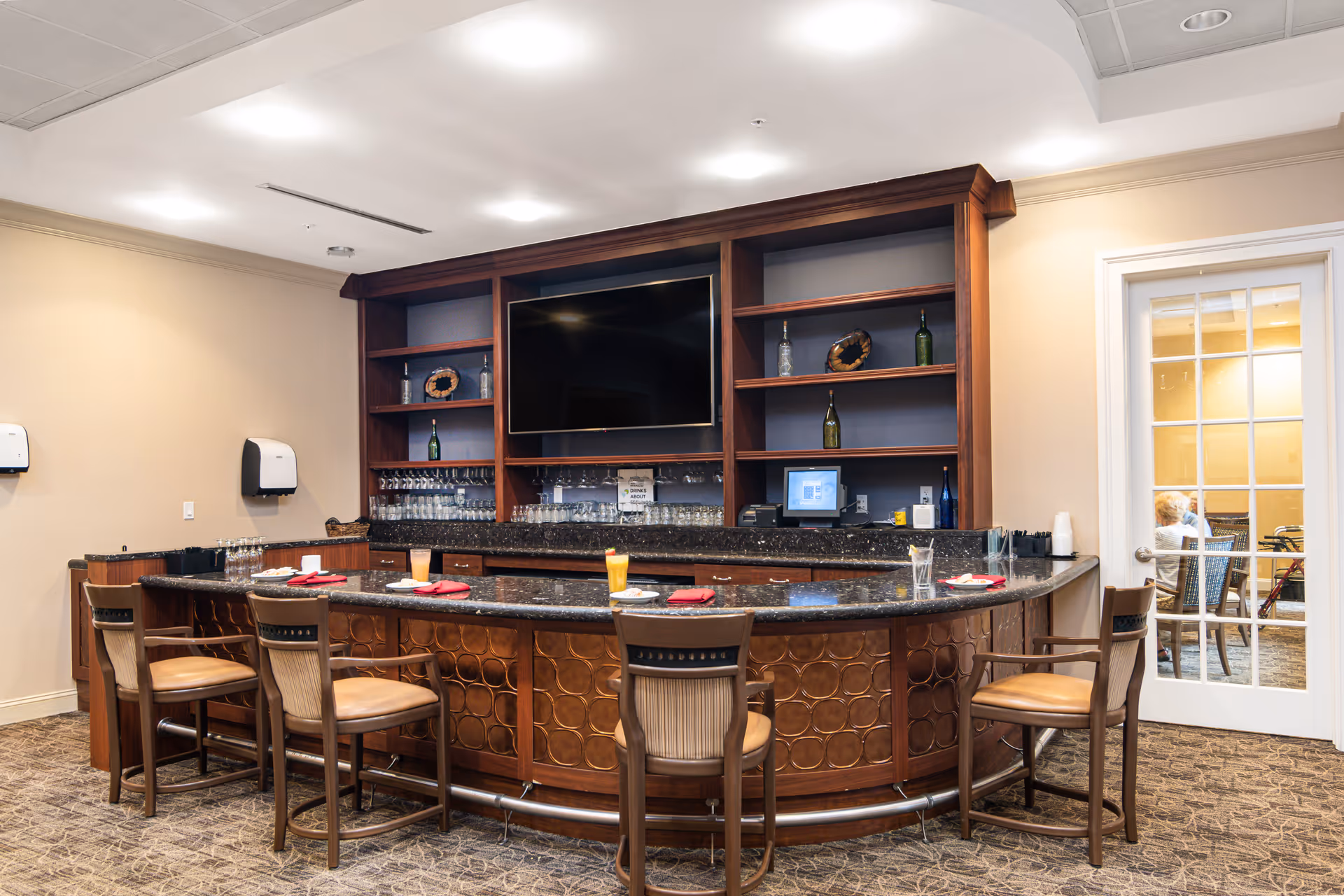 Interior view of a senior living facility bar area with a curved wooden counter, five wooden chairs with cushioned seats, a large flat-screen TV mounted on a wooden shelving unit behind the bar, and various bottles and glassware on the shelves. There is a glass door on the right side showing a glimpse of another room with people seated.