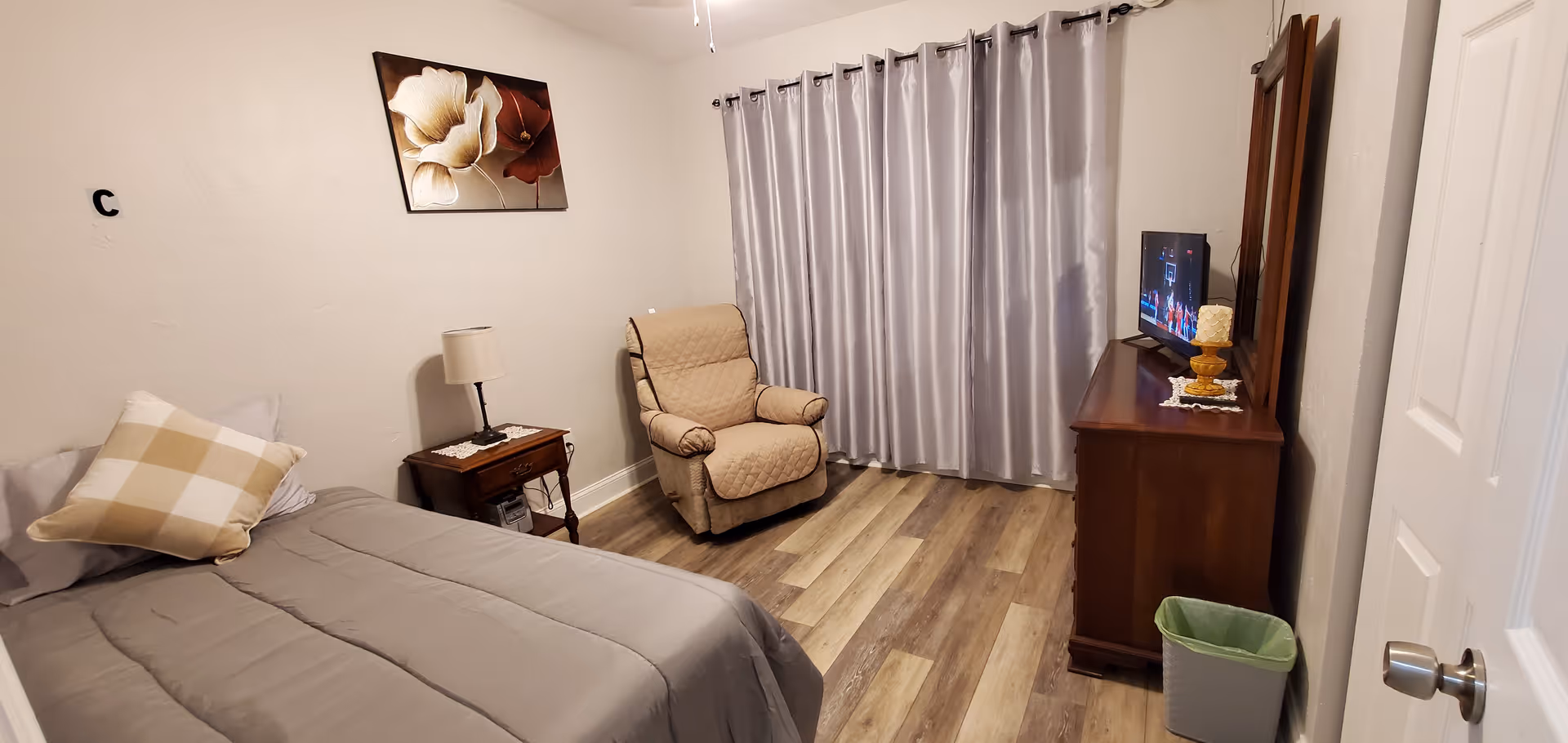 A cozy bedroom with a bed covered in gray bedding and a checkered pillow, a wooden nightstand with a lamp, a beige recliner chair, a wooden dresser with a TV on top, a green trash bin, and gray curtains covering a window. The walls are light-colored and there is a floral painting hanging above the nightstand.