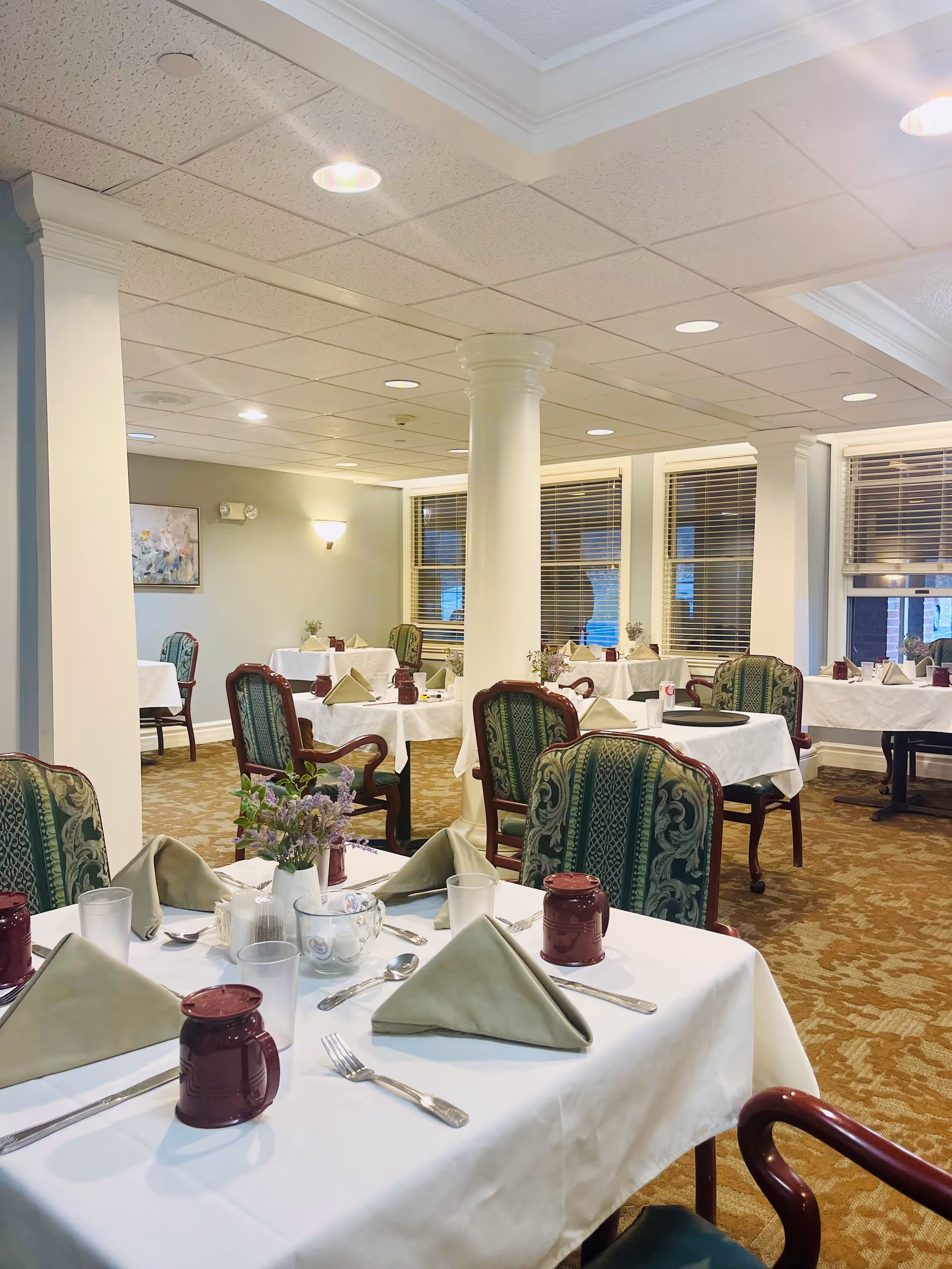 A dining room in a senior living facility with tables covered in white tablecloths, each set with folded beige napkins, silverware, clear drinking glasses, and maroon mugs. The room features green upholstered chairs with wooden frames, beige patterned carpet, white columns, and windows with blinds. Soft lighting illuminates the space, creating a warm and inviting atmosphere.