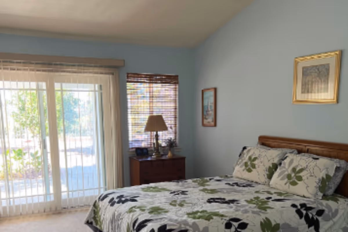 A bedroom with a bed covered in a floral patterned bedspread in shades of green, black, and gray. There is a wooden nightstand with a lamp and some decorative items next to a window with bamboo blinds. Sliding glass doors with vertical blinds lead outside, allowing natural light to fill the room. The walls are painted light blue and have framed artwork hanging on them.
