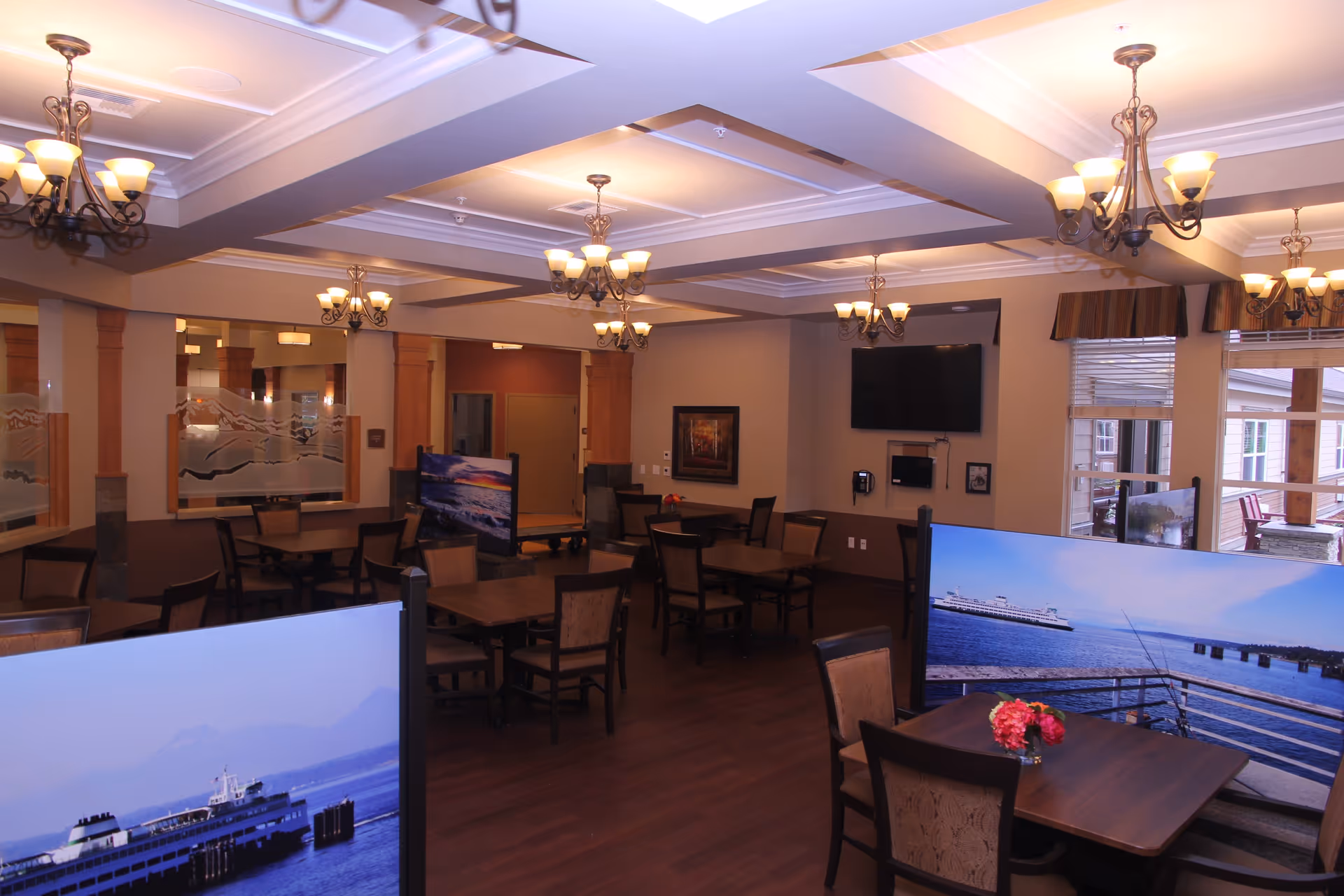 A spacious dining room with multiple wooden tables and chairs arranged neatly. The room features warm lighting from several chandeliers hanging from a coffered ceiling. Decorative panels with scenic ocean views separate some tables. A large flat-screen TV is mounted on one wall, and windows with blinds allow natural light to enter. The overall ambiance is cozy and inviting.