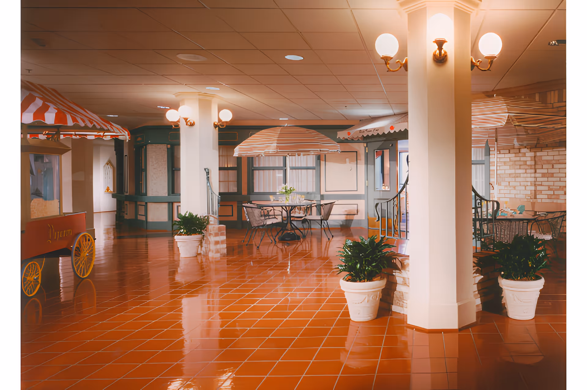 Indoor common area with shiny terracotta tile flooring, white pillars with wall-mounted globe lights, potted plants, and tables with striped umbrellas. There is a red popcorn cart on the left side and a cozy seating area in the background.