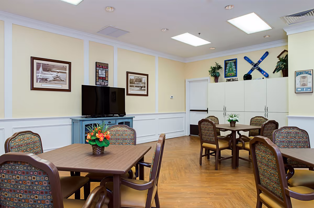 A well-lit common room with several wooden tables and cushioned chairs arranged around them. A flat-screen TV sits on a blue cabinet against a wall decorated with framed pictures and sports-themed posters. The room has light yellow walls with white wainscoting and a wood-patterned floor. Potted plants and floral centerpieces add a touch of greenery to the space.