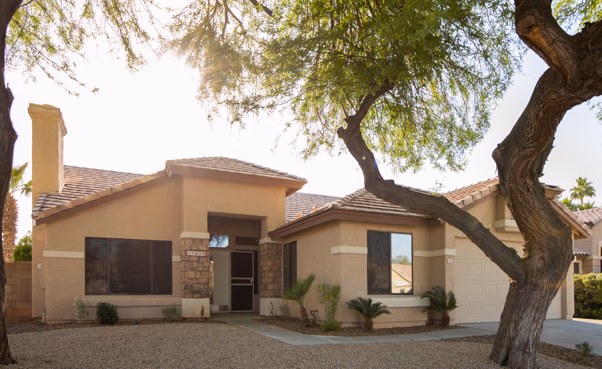 Exterior view of a single-story beige stucco house with a tiled roof, a large tree in the front yard, and a driveway leading to a garage. The yard is landscaped with gravel and small plants.