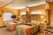 Shared senior-care bedroom with two twin beds, bedside tables, a wall-mounted TV, and warm yellow-orange walls.