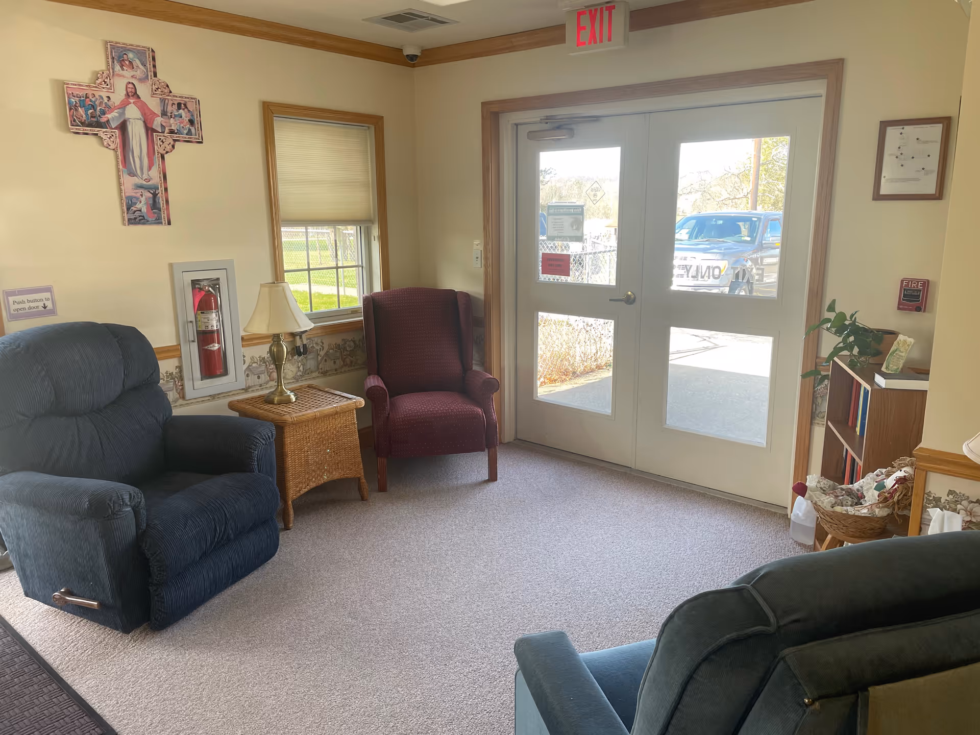 A small seating area near the exit doors of a facility, featuring two upholstered armchairs, a wicker side table with a lamp, a window with blinds, a fire extinguisher mounted on the wall, a religious cross decoration, and a small bookshelf with a plant and other items.
