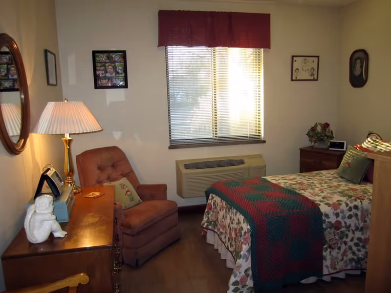 A cozy bedroom with a floral bedspread and a colorful quilt on the bed. There is a wooden dresser with a lamp, a small statue, and a radio on it. A cushioned armchair with a decorative pillow is placed next to the dresser. The room has a window with blinds and a red valance, and framed pictures hang on the walls.