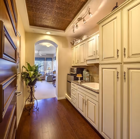 A narrow kitchen area with cream-colored cabinets and drawers on the right side, a countertop with a sink, a microwave, and decorative items. The floor is dark wood, and there is a potted plant on a decorative stand on the left side near a wooden door. The kitchen opens into a living room area with large windows and seating visible in the background.