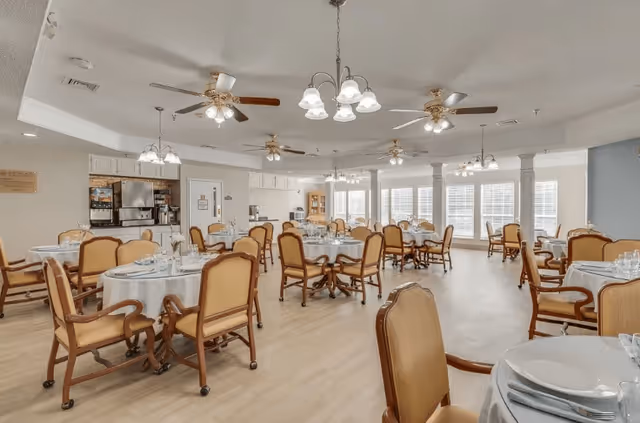 A spacious dining room with multiple round tables covered with white tablecloths, each set with plates, glasses, and silverware. The room features wooden chairs with cushioned seats and backs, ceiling fans, and hanging light fixtures. Large windows along one wall allow natural light to fill the space. A kitchen area with stainless steel appliances is visible in the background.