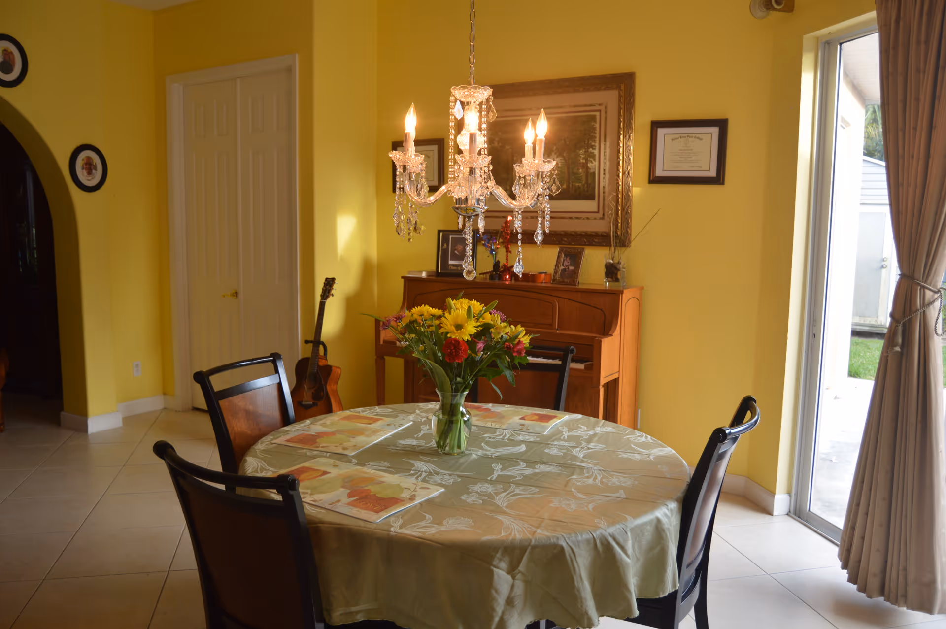 A dining room with yellow walls featuring a round table covered with a floral tablecloth and a vase of colorful flowers in the center. Four chairs surround the table. A crystal chandelier hangs above the table. In the background, there is a wooden piano with framed pictures and a certificate on the wall above it. A guitar leans against the wall near a closed door. To the right, a sliding glass door with beige curtains opens to an outdoor area.
