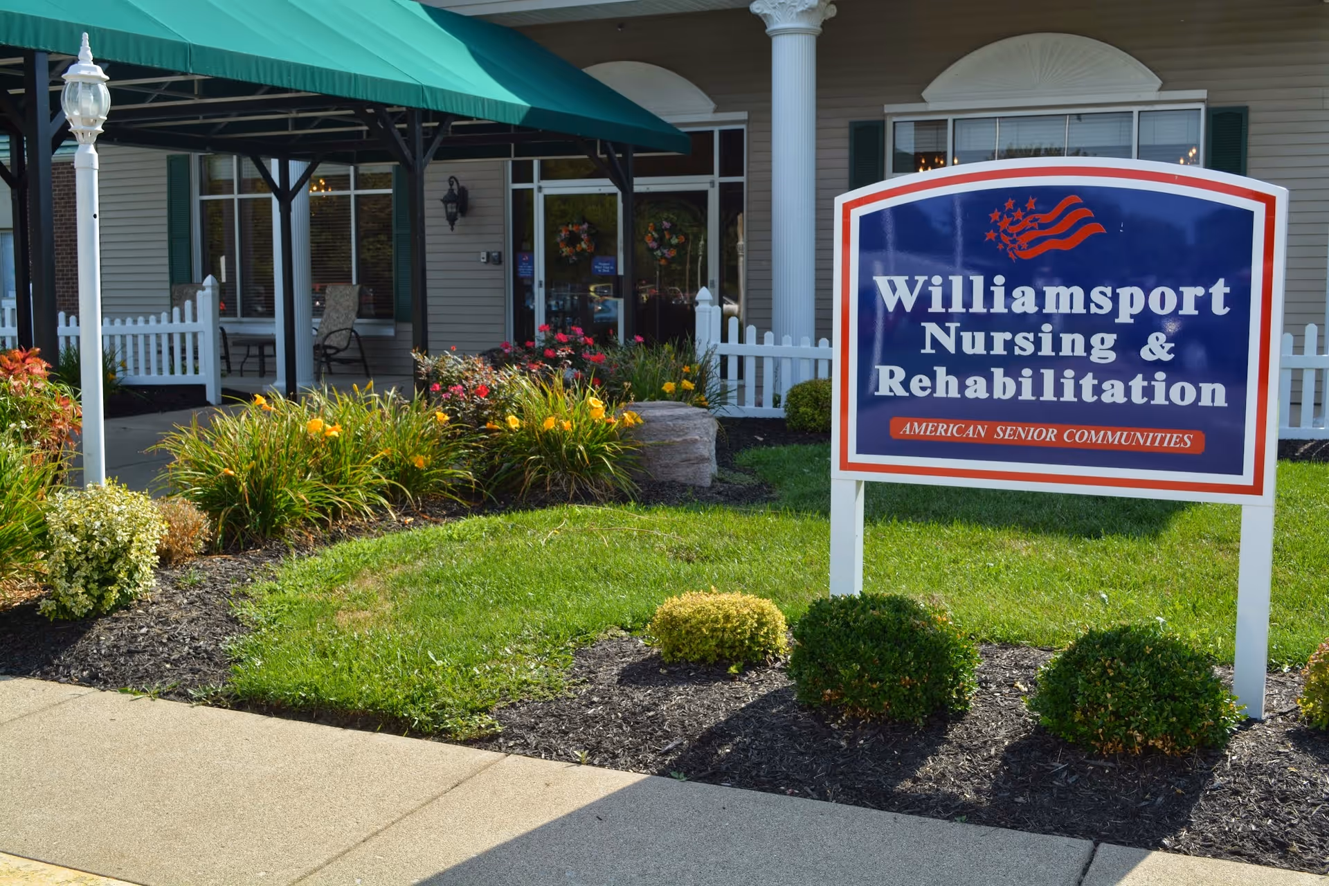 Front entrance of Williamsport Nursing & Rehabilitation with a large blue sign, landscaped lawn, and covered walkway.
