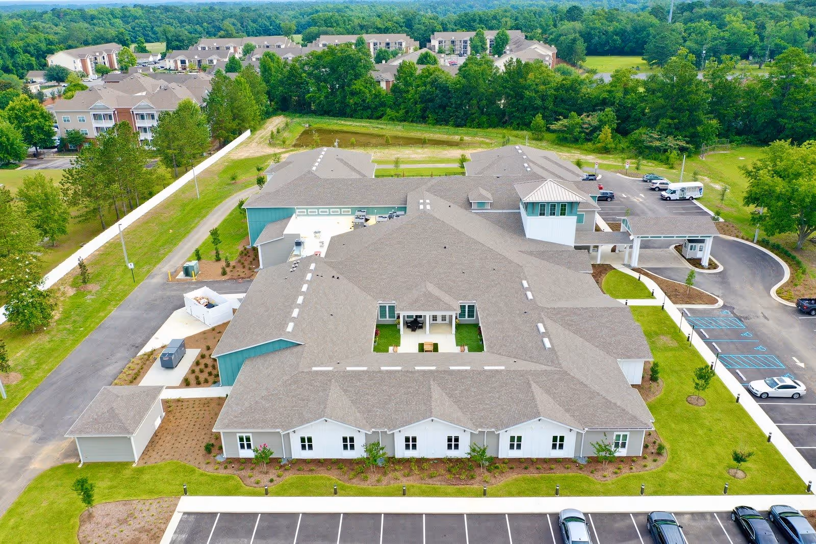 Aerial view of The Canopy at Azalea Grove senior living facility showing a large building with multiple roof sections, surrounded by green lawns, parking lots, and trees. Residential buildings and wooded areas are visible in the background.
