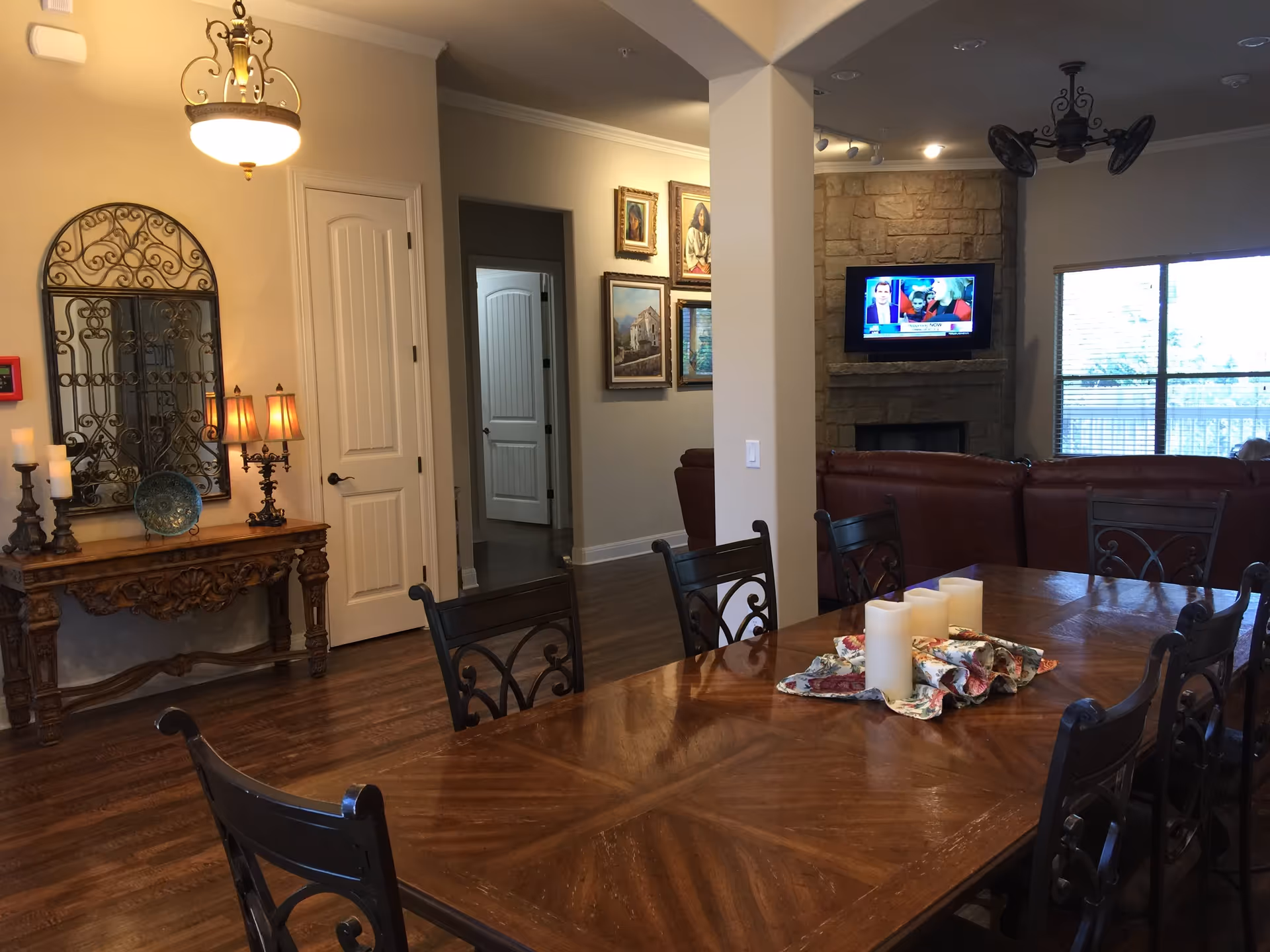 Interior view of a living and dining area in an assisted living facility. The foreground features a large wooden dining table with decorative candles and a floral cloth centerpiece. Surrounding the table are several dark metal chairs with intricate designs. In the background, there is a stone fireplace with a mounted flat-screen TV above it, showing a news broadcast. To the left, a wooden console table with candles, a decorative plate, and a lamp sits beneath a large ornate mirror. The room has hardwood floors, beige walls, and several framed paintings on the walls. Large windows on the right side allow natural light to enter the space.