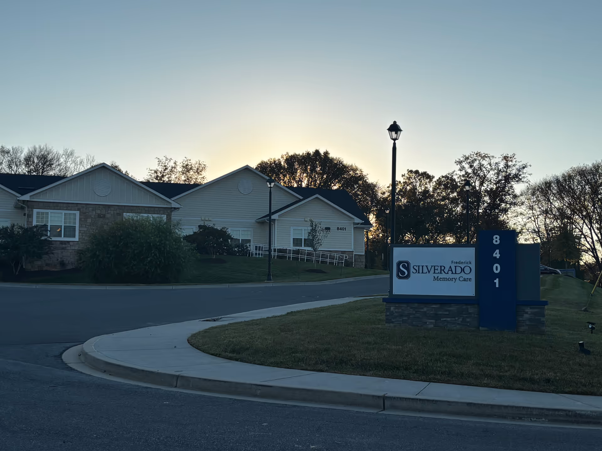 Exterior view of Silverado Frederick Memory Care Community building at sunset with a sign displaying the facility name and address number 8401, surrounded by trees and a curved driveway.