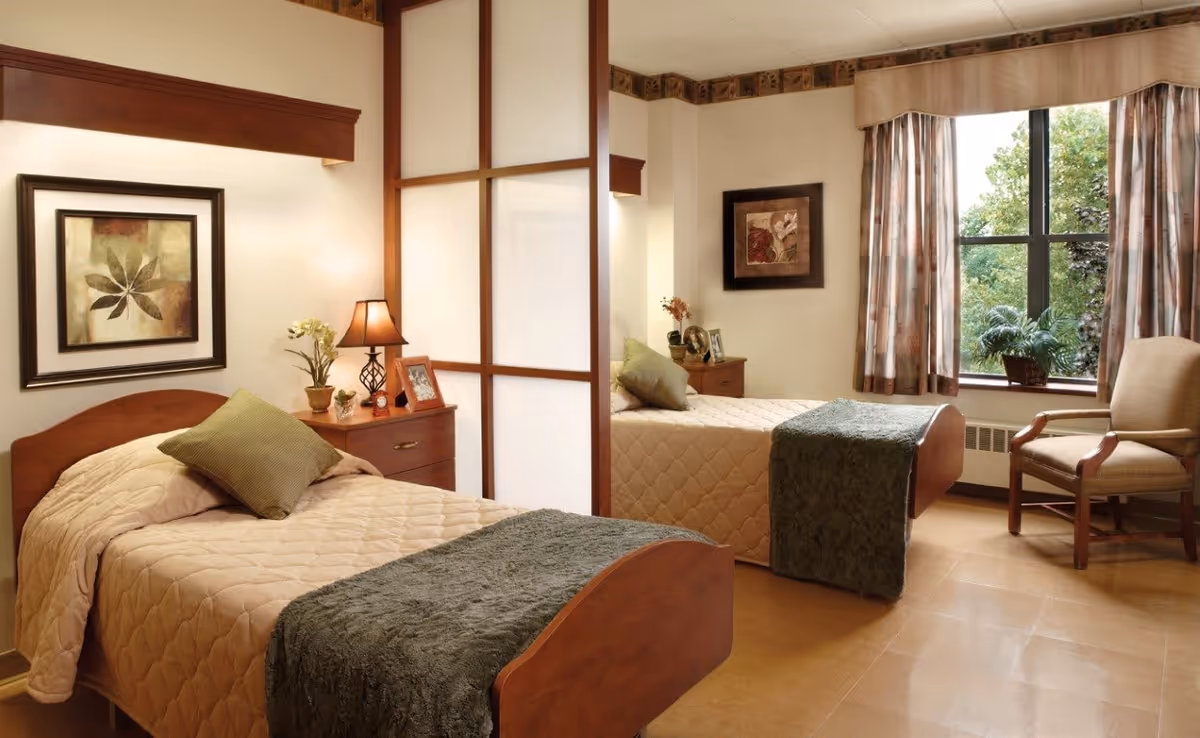 A cozy bedroom in a senior living facility with two single beds separated by a wooden and frosted glass partition. Each bed has beige quilted bedding, a green throw blanket, and a green pillow. There are wooden nightstands with lamps, framed photos, and small plants. A large window with patterned curtains lets in natural light, and a cushioned armchair is placed near the window. The walls are decorated with framed artwork.