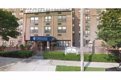 Exterior view of Throgs Neck Rehabilitation & Nursing Center, a multi-story brick building with a blue awning over the main entrance. There are trees and shrubs along the sidewalk and a sign near the entrance.