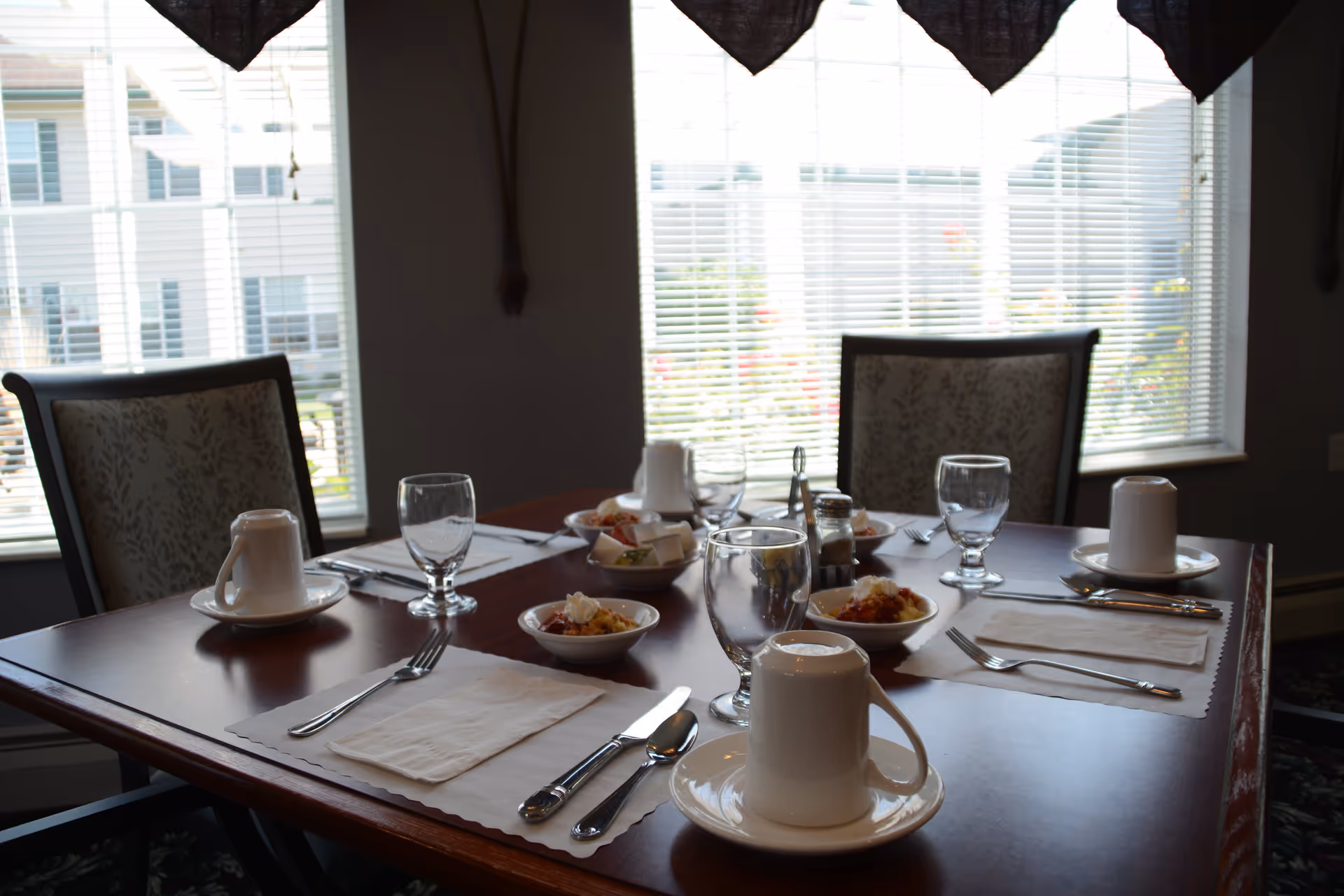 A dining table set for four with white placemats, silverware, upside-down white coffee cups on saucers, empty water glasses, and small bowls of food. The table is near large windows with blinds, allowing natural light to enter the room. Upholstered chairs with floral patterns surround the table.