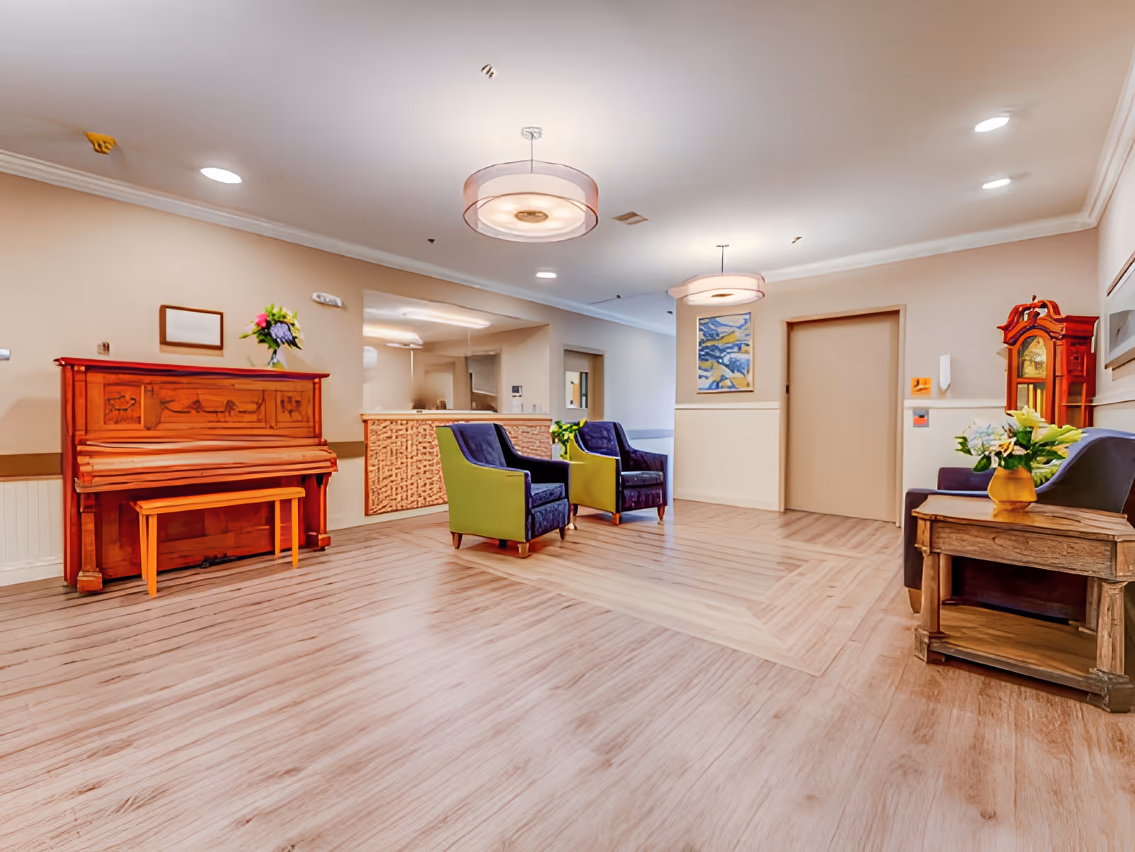 A well-lit interior common area with light wood flooring, beige walls, and ceiling lights. The room features a wooden upright piano with a bench and a flower vase on top, two blue and green armchairs, a wooden side table with a flower vase, and a tall wooden grandfather clock. There is a reception desk in the background and a painting on the wall near a closed door.
