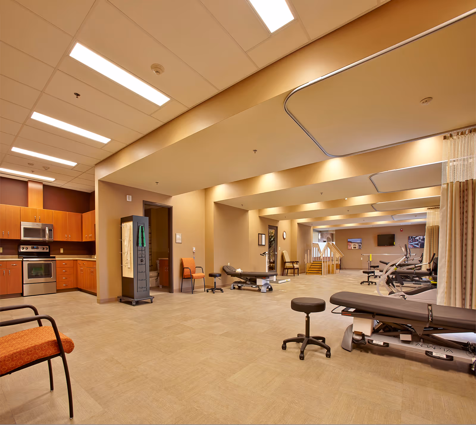 A spacious rehabilitation or physical therapy room with exercise equipment including therapy tables, stools, and exercise machines. The room has warm beige walls, overhead fluorescent lighting, and a kitchen area with wooden cabinets and stainless steel appliances on the left side. There are chairs placed around the room and a small set of stairs in the background.