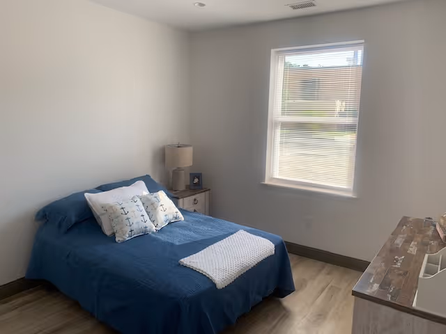 Simple bedroom with a blue-covered bed, nightstand and lamp, a dresser, and a window letting in daylight.