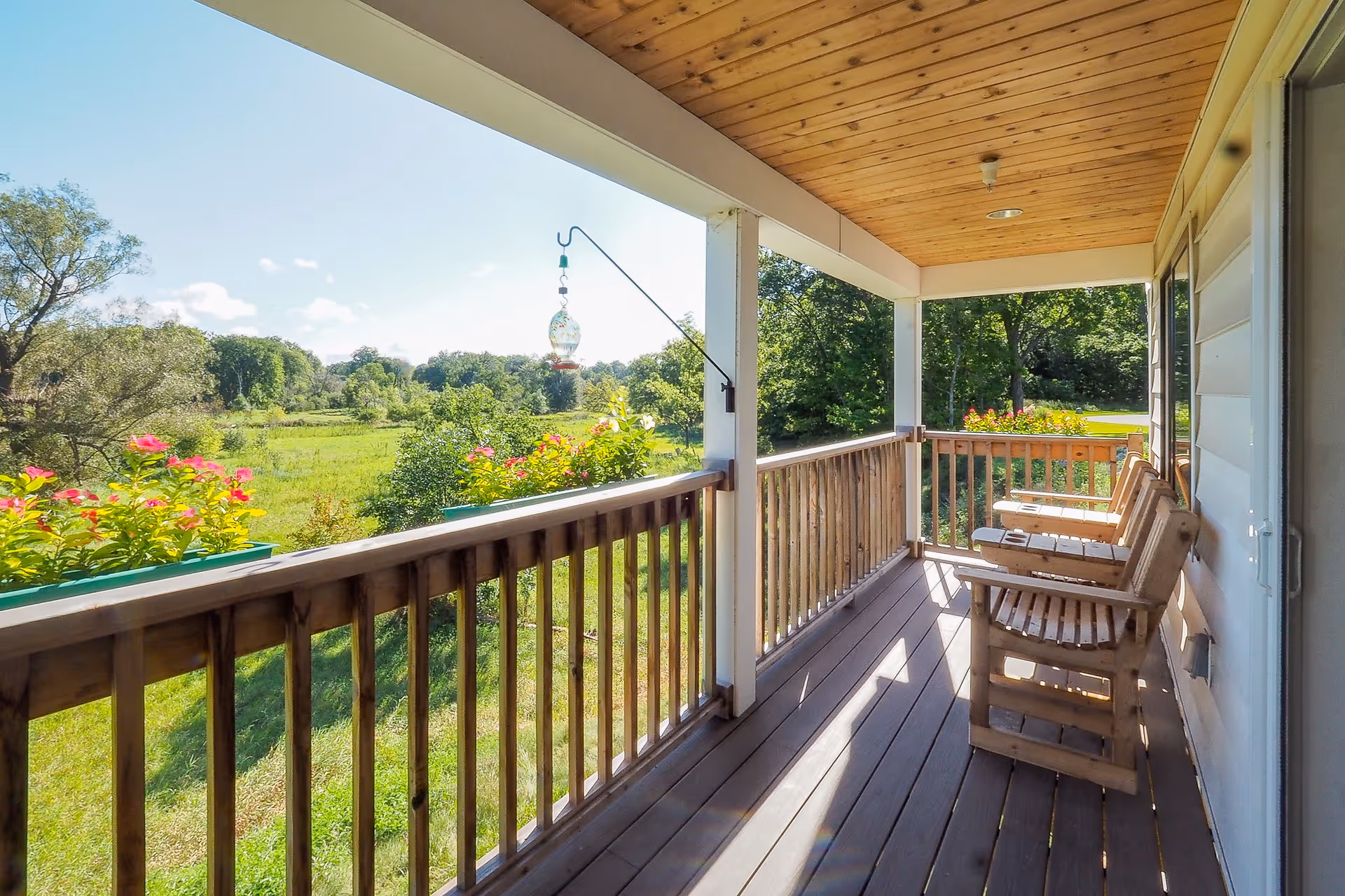 A covered wooden porch with three wooden chairs facing a scenic view of green trees and a grassy field. Flower boxes with pink flowers are attached to the porch railing, and a hanging bird feeder is visible. The porch ceiling is made of wood panels.