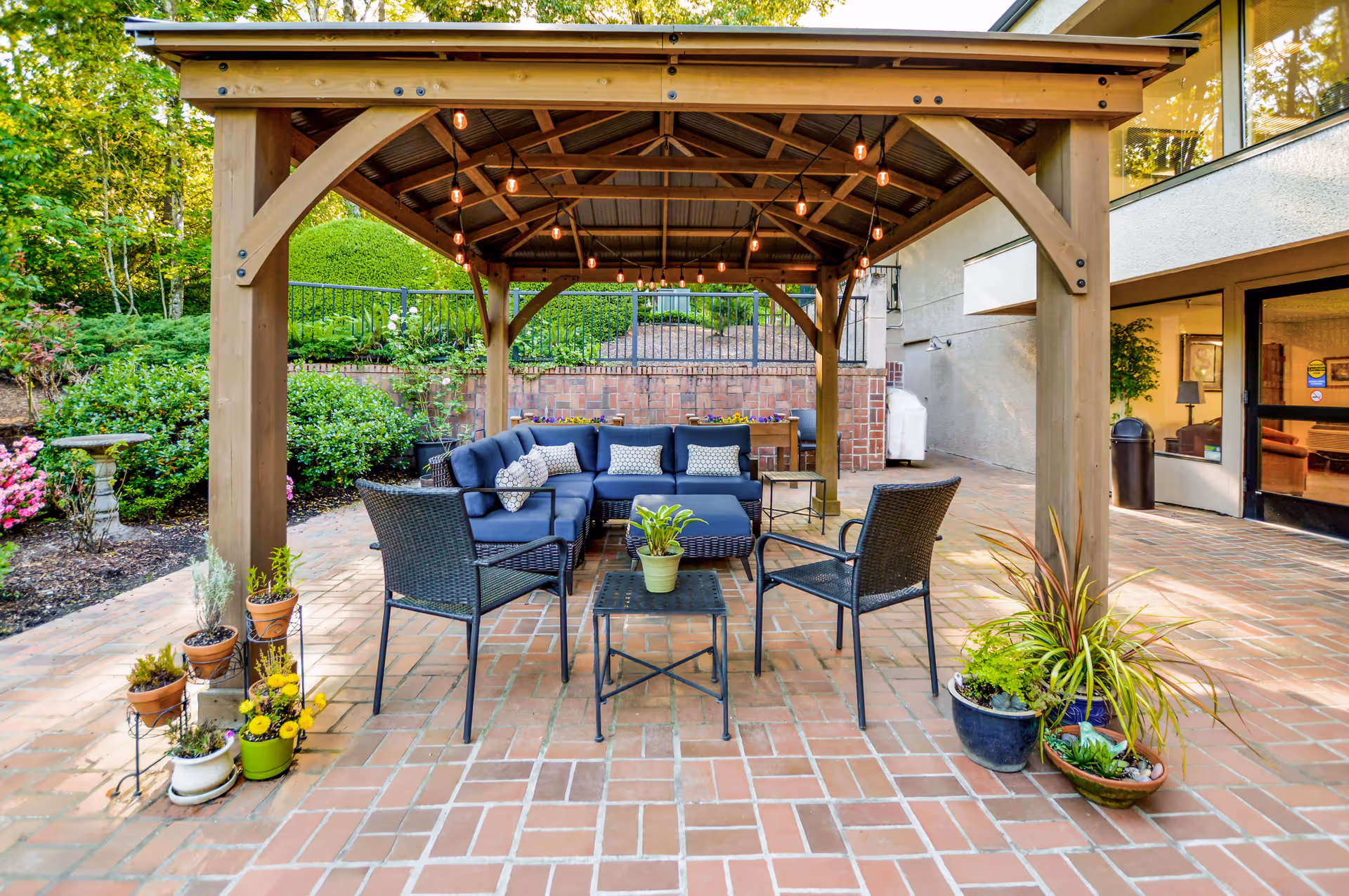 Outdoor patio area with a wooden pergola overhead, string lights hanging from the roof, a blue cushioned sectional sofa, two black wicker chairs, and a small table with a potted plant. Surrounding the patio are various potted plants and greenery, with a brick wall and building entrance visible in the background.