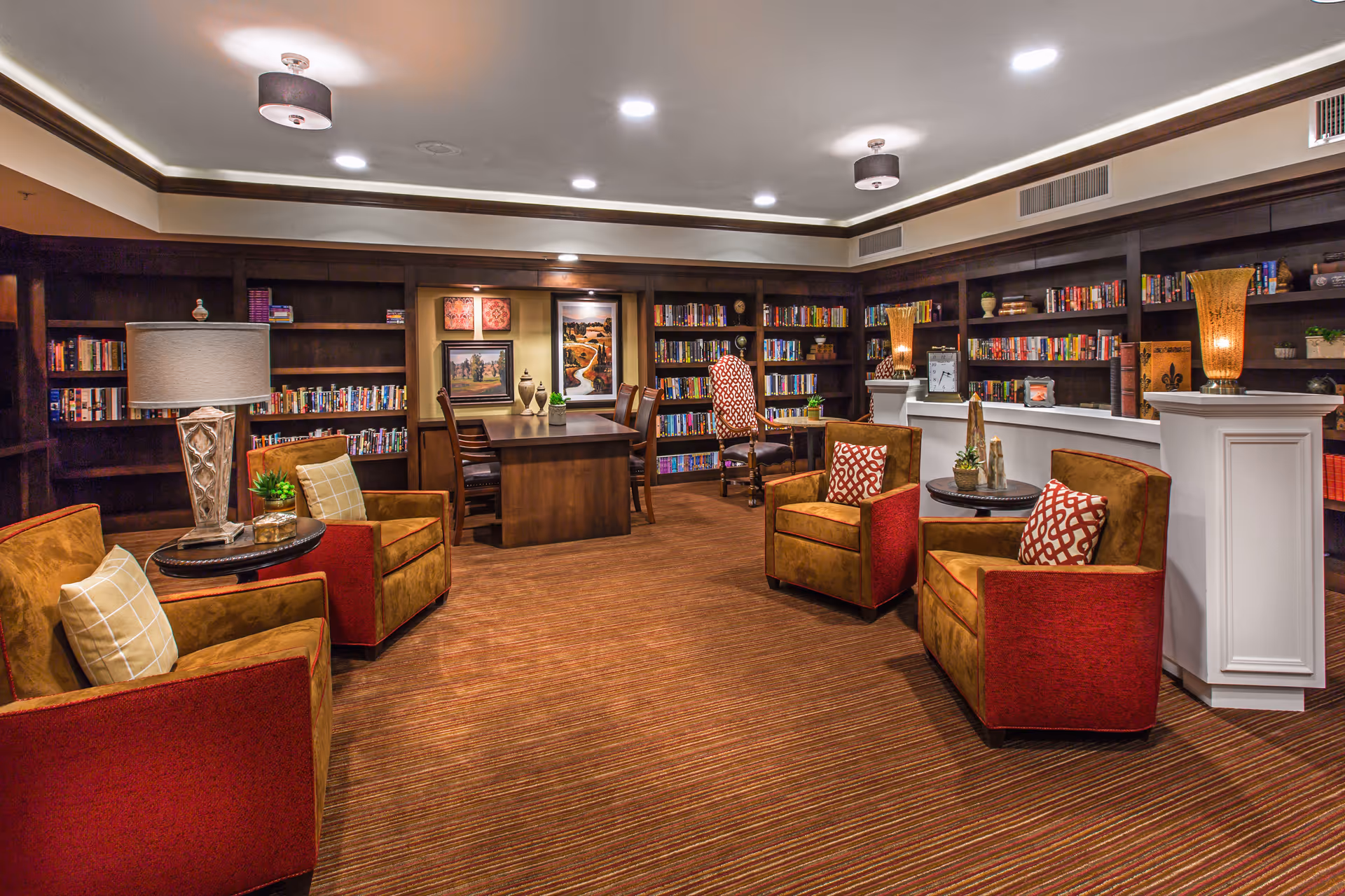 Cozy library-style lounge with bookshelves, upholstered armchairs, lamps, and a central table.