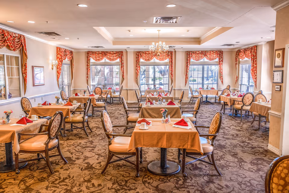 A bright and elegant dining room with multiple tables covered in beige tablecloths, each set with red folded napkins, white cups, and small flower vases. The room features patterned carpet, large windows with red and gold curtains, and a chandelier hanging from the ceiling.