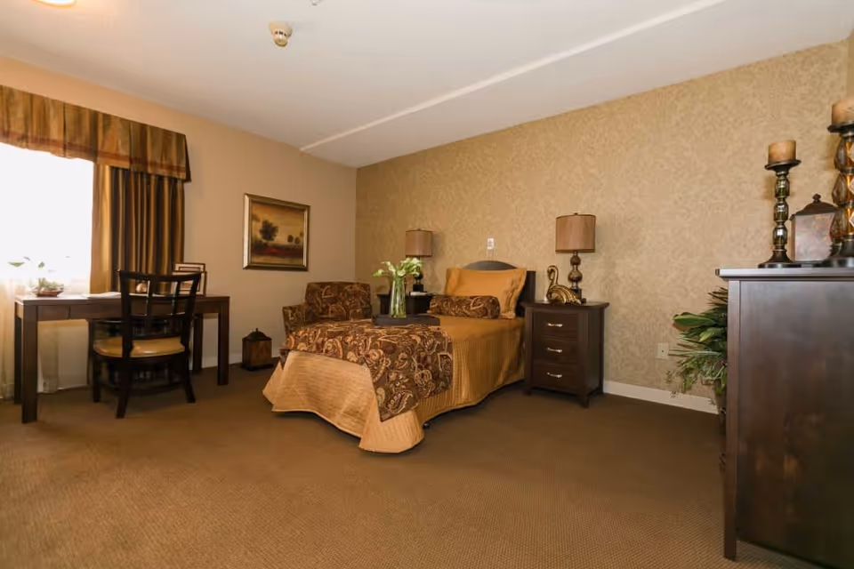 A cozy senior living bedroom with a single bed covered in a brown and gold patterned bedspread. Next to the bed is a dark wooden nightstand with a lamp and decorative items. There is a matching armchair with a similar pattern to the bedspread. A wooden desk with a chair is positioned near a window with brown curtains. The walls are beige with a subtle patterned wallpaper on one side, and the floor is carpeted in brown.