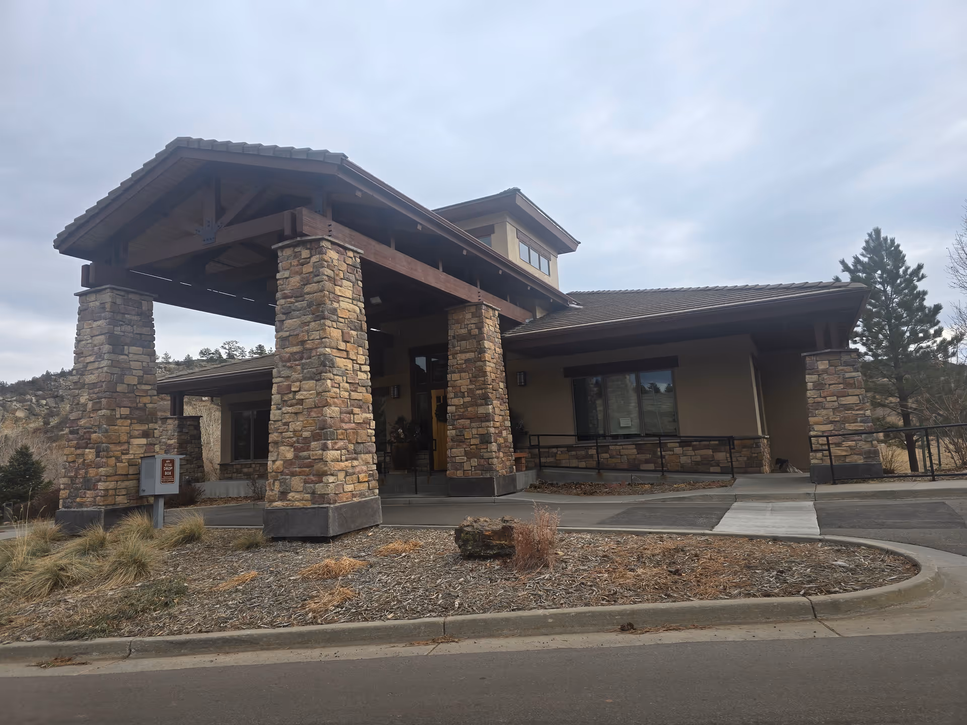 Exterior view of a building with a covered entrance supported by stone pillars, surrounded by dry landscaping and trees under a cloudy sky.