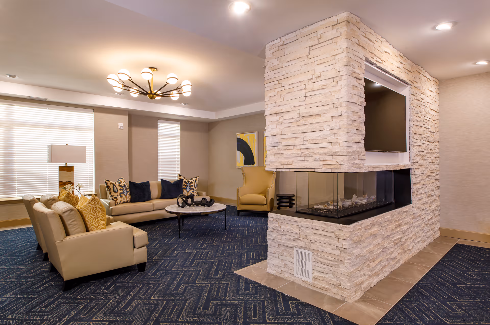 A modern living room area in an assisted living facility featuring beige leather sofas with decorative pillows, a round coffee table, a yellow armchair, and a central stone fireplace with a built-in TV. The room has large windows with blinds, a contemporary ceiling light fixture, and blue patterned carpet.