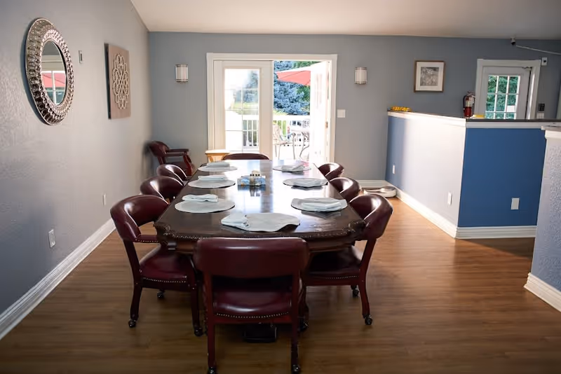 A dining room with a long wooden table set with placemats and napkins, surrounded by maroon chairs. The room has light blue walls, a wooden floor, a decorative round mirror on the left wall, and double glass doors leading to an outdoor patio with an umbrella.