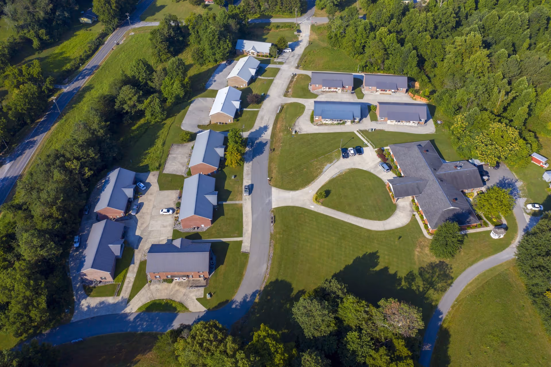 Aerial view of Small Town Senior Living at Deer Ridge showing multiple single-story buildings with gray roofs arranged along paved roads and surrounded by green lawns and trees.