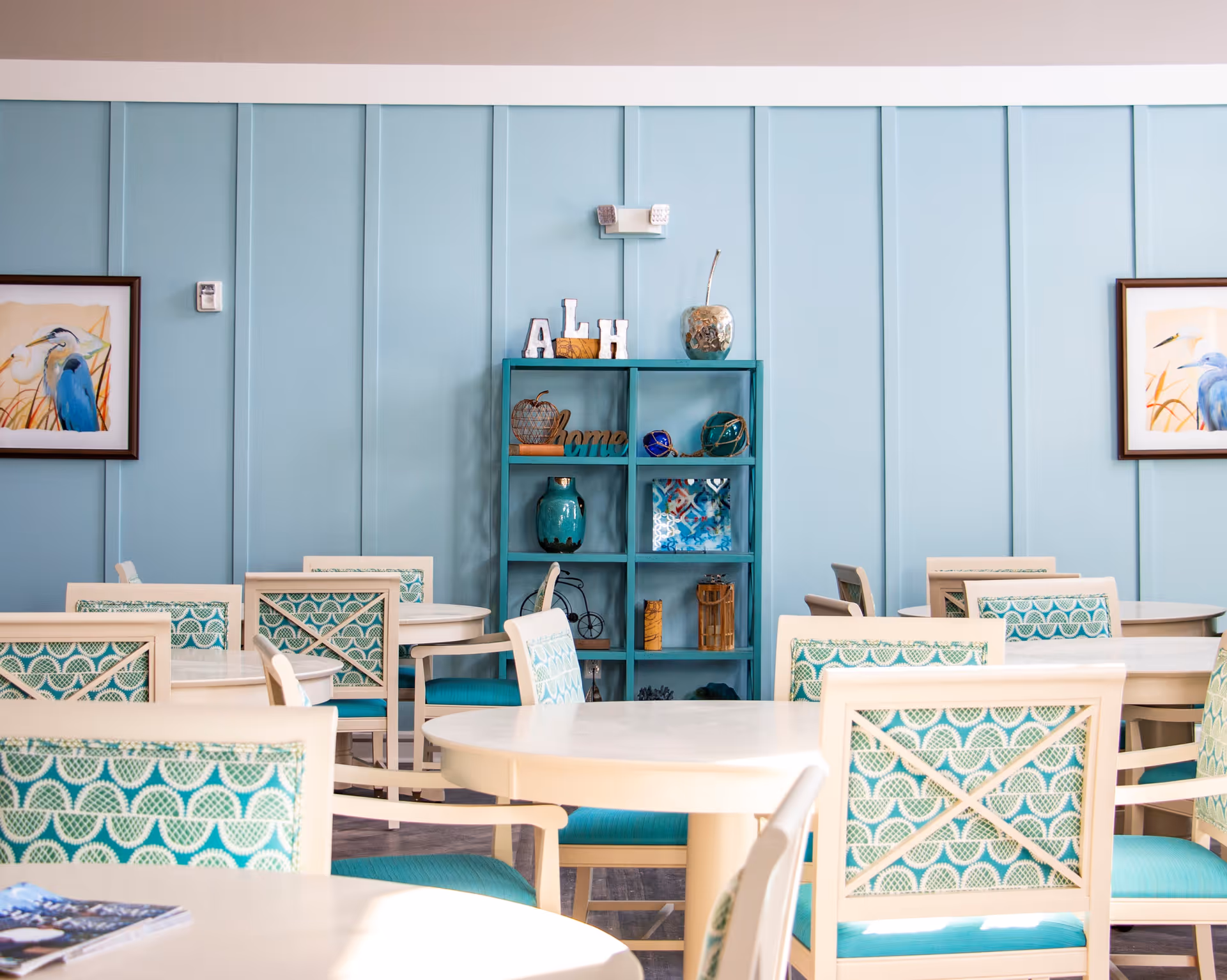 A bright dining area with round white tables and chairs featuring teal and white patterned cushions. The walls are painted light blue with vertical paneling. A teal shelving unit with decorative items is centered on the back wall, flanked by framed bird artwork on either side.