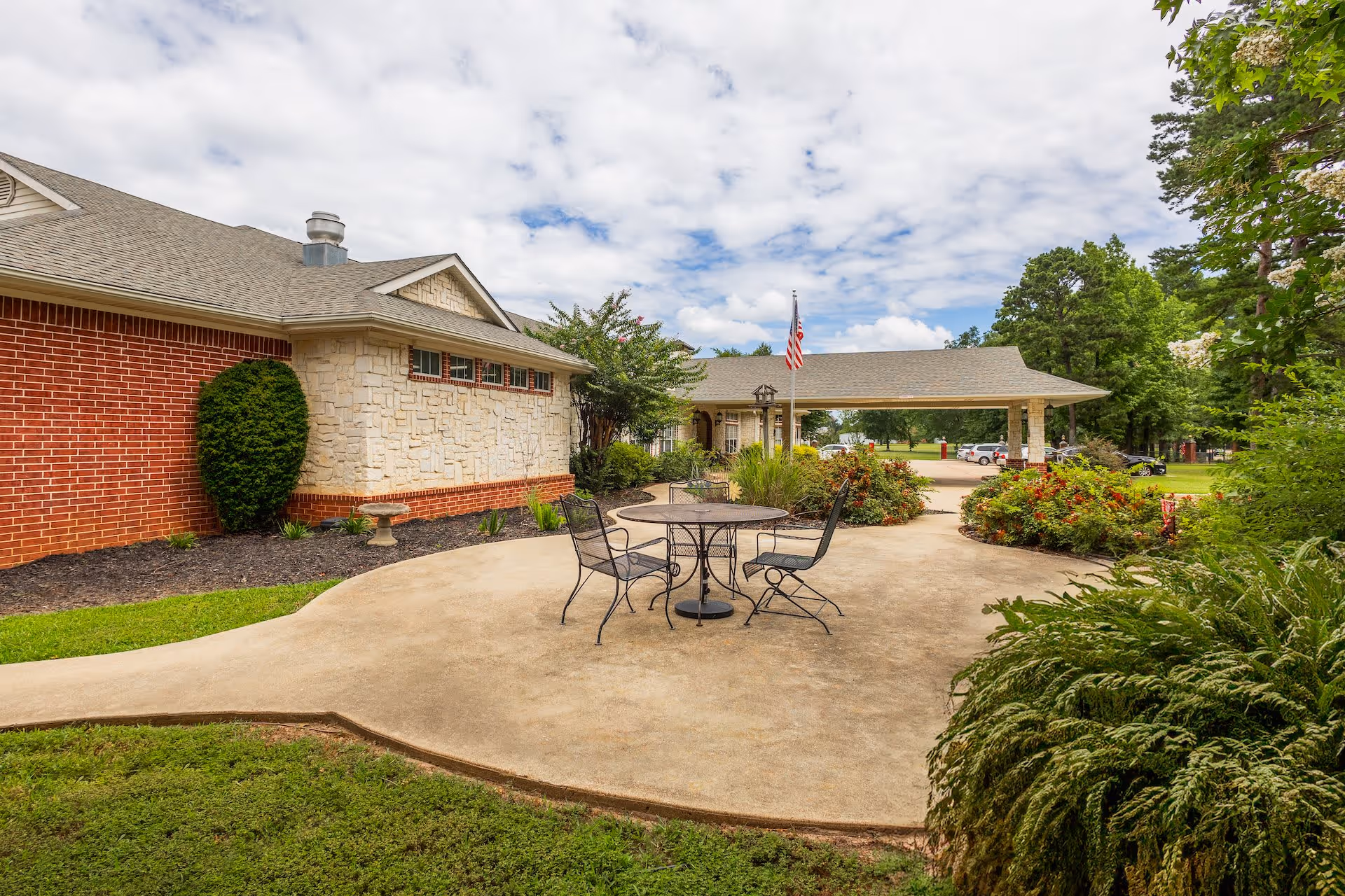 Outdoor patio area at a senior living facility with a round metal table and four chairs on a concrete surface, surrounded by greenery and bushes. The building has a brick and stone exterior with a covered entrance and an American flag flying near the entrance. The sky is partly cloudy.