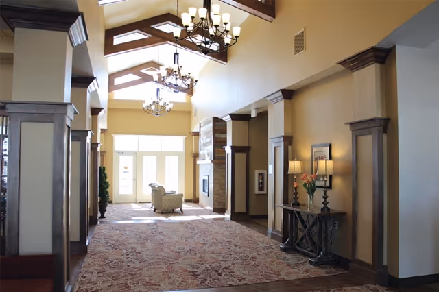 A spacious and well-lit interior hallway with high ceilings featuring exposed wooden beams and chandeliers. The hallway has a patterned carpet and cream-colored walls with dark wood trim. At the far end, there are large glass doors letting in natural light, and a cozy seating area with an armchair near a stone fireplace. A console table with lamps and flowers is positioned along one wall.