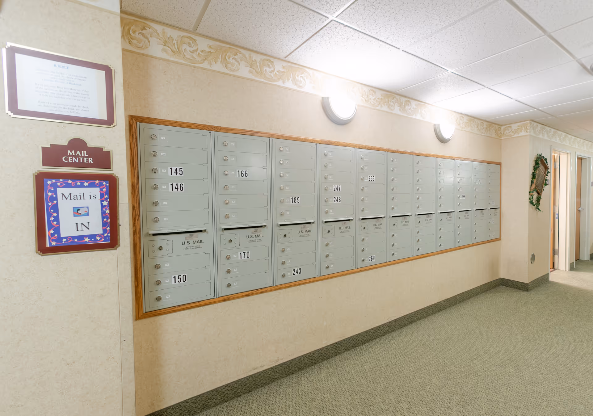 A hallway in a senior living facility with a wall-mounted mail center featuring multiple locked mailboxes labeled with numbers. There is a sign next to the mailboxes that reads 'Mail is IN'. The hallway has beige walls with a decorative border near the ceiling, green carpet, and ceiling lights.
